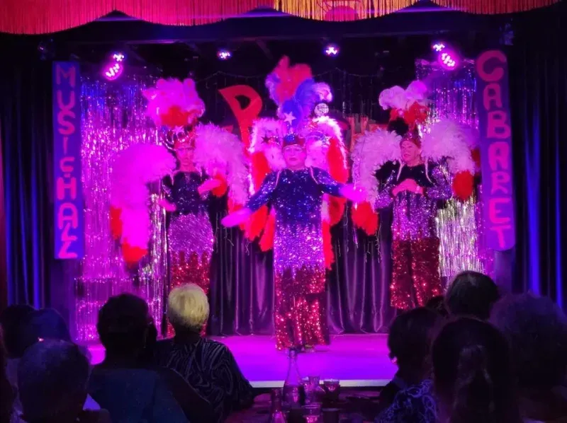 La troupe sur la scène du Cabaret Chez Bibiche, en costumes à plumes, devant le public.